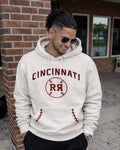 Man wearing a white hoodie with 'Cincinnati RR' print, standing in front of a brick building.