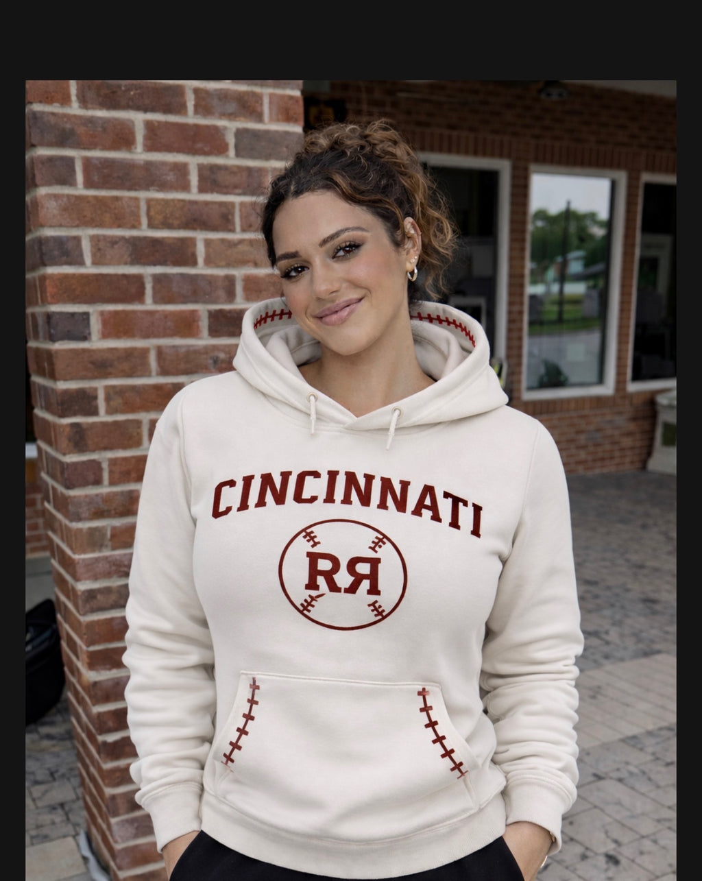 Woman wearing a white hoodie with 'Cincinnati' and a logo, standing against a brick wall.